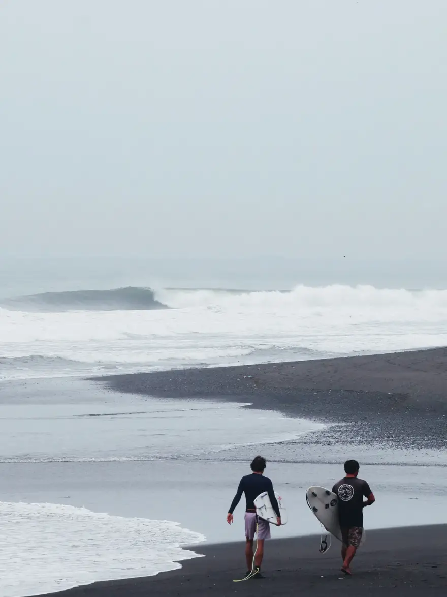 A surf coach and a surfer walking down a black sand beach in Bali towards a clean peeling wave