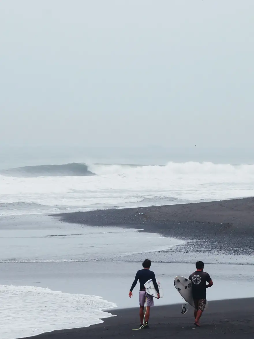 Two surfers walking down a black sand beach in Bali towards a clean peeling wave