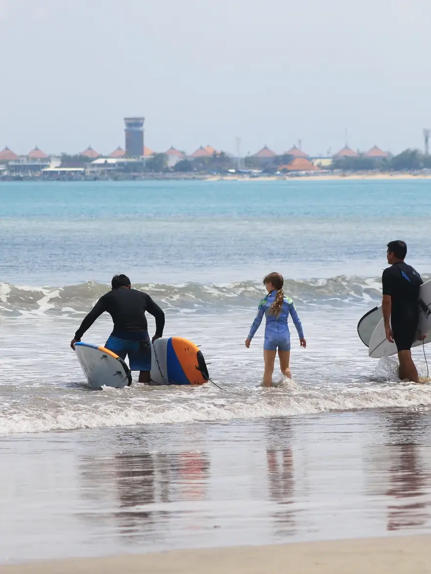 A surf instructor walking a young girl and her surfboard into small waves on a Bali beach